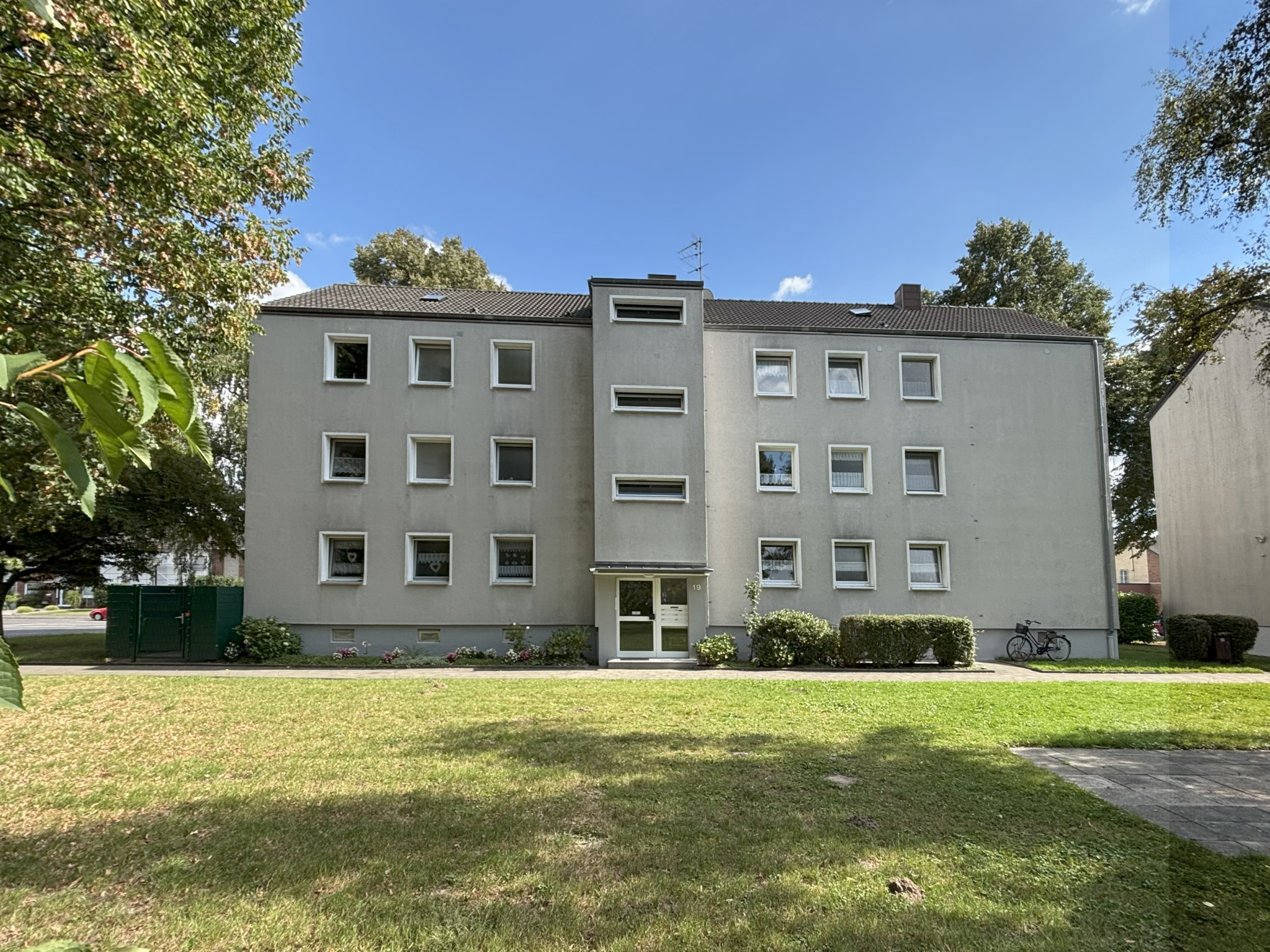 Das Bild zeigt die Außenansicht eines dreigeschossigen Mehrfamilienhauses mit schlichtem, hellgrauem Putz und Satteldach.