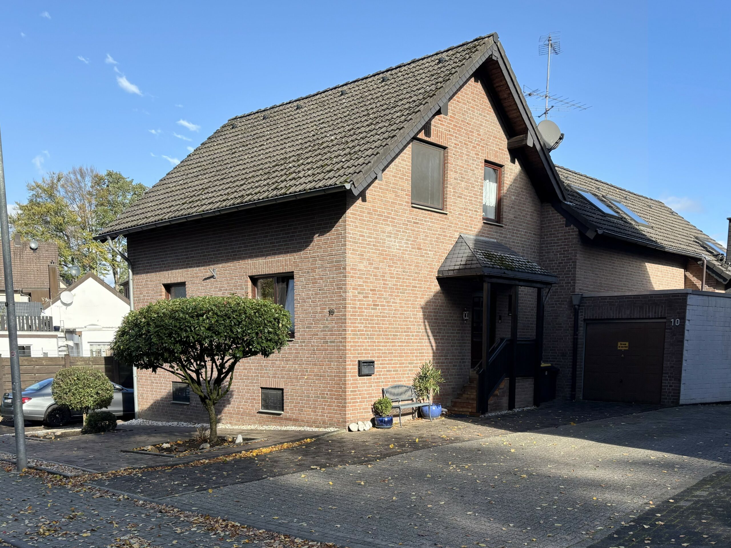 Das Bild zeigt den Eingangsbereich eines Einfamilienhauses. Ein gepflasterter Vorgarten mit zwei Sträuchern und die Einfahrt zur Garage sind zu sehen.