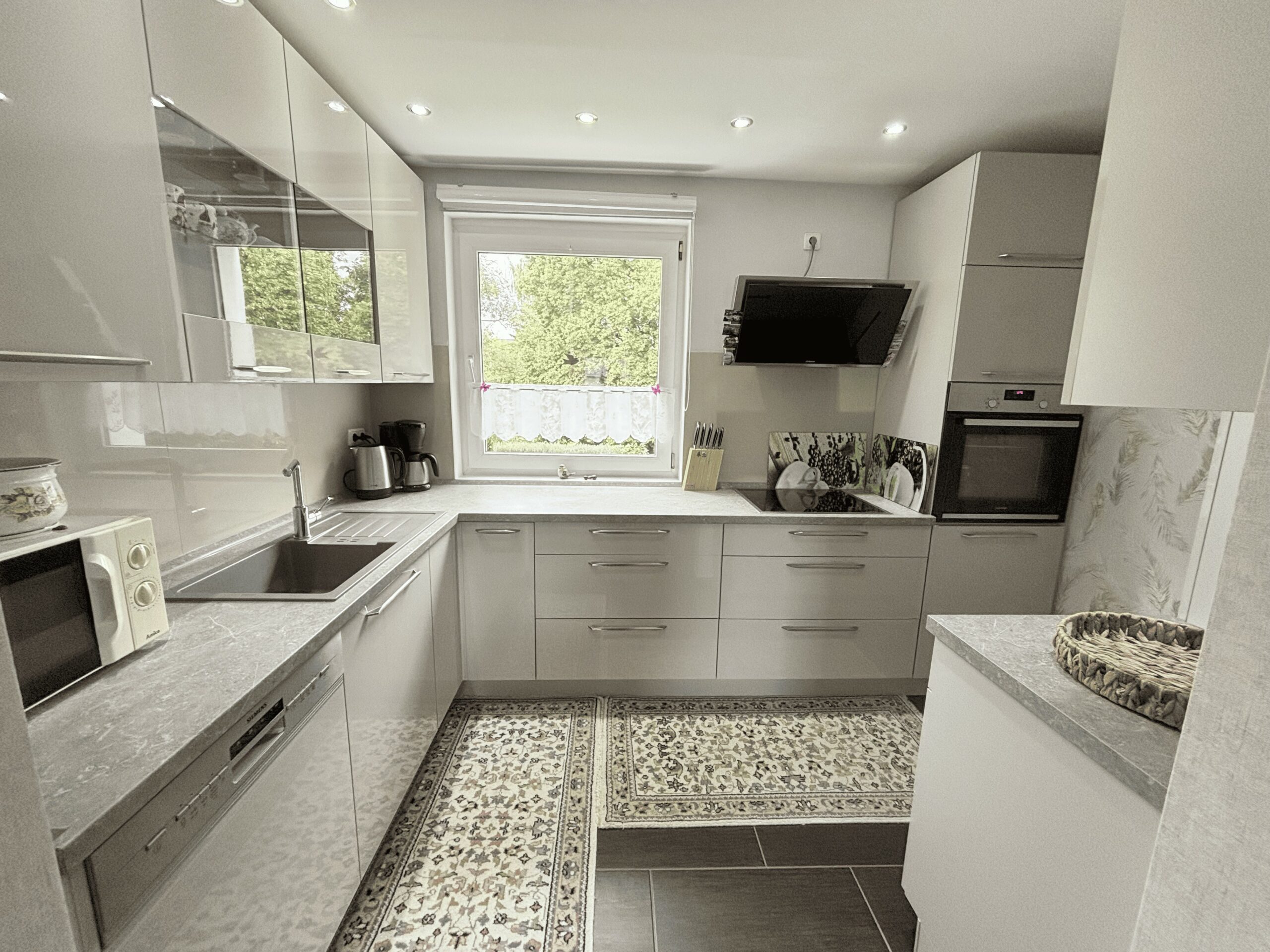 Das Bild zeigt eine moderne, helle Einbauküche in U-Form mit Hochglanz-Fronten. Über dem Spülbecken befindet sich ein Fenster.