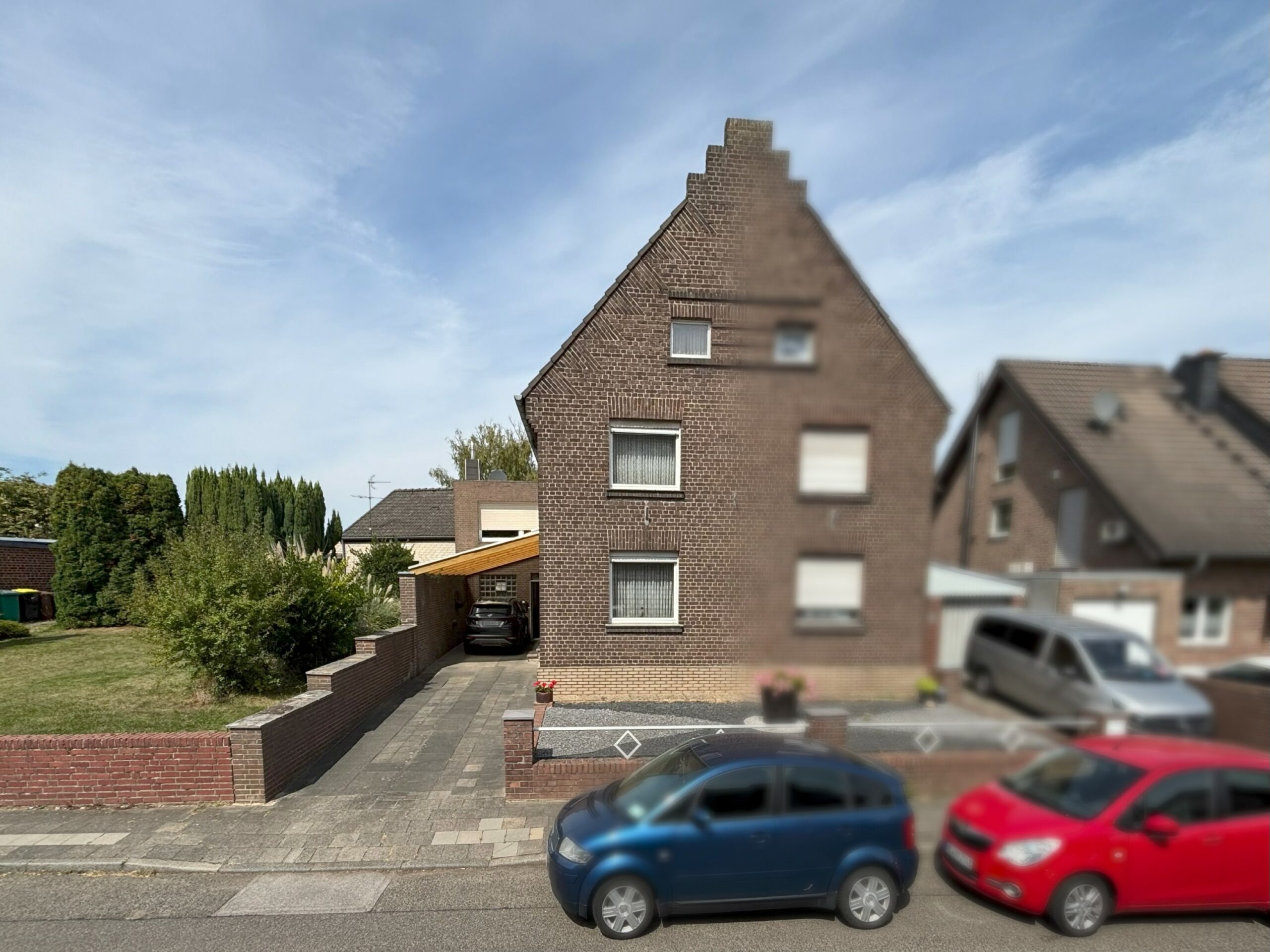 Das Bild zeigt eine zweieinhalbgeschossigDoppelhaushälfte aus dunklem Klinker mit Satteldach und Treppengiebel. Vor dem Haus stehen Autos am Straßenrand, rechts führt eine Einfahrt mit Carport zum Garten.