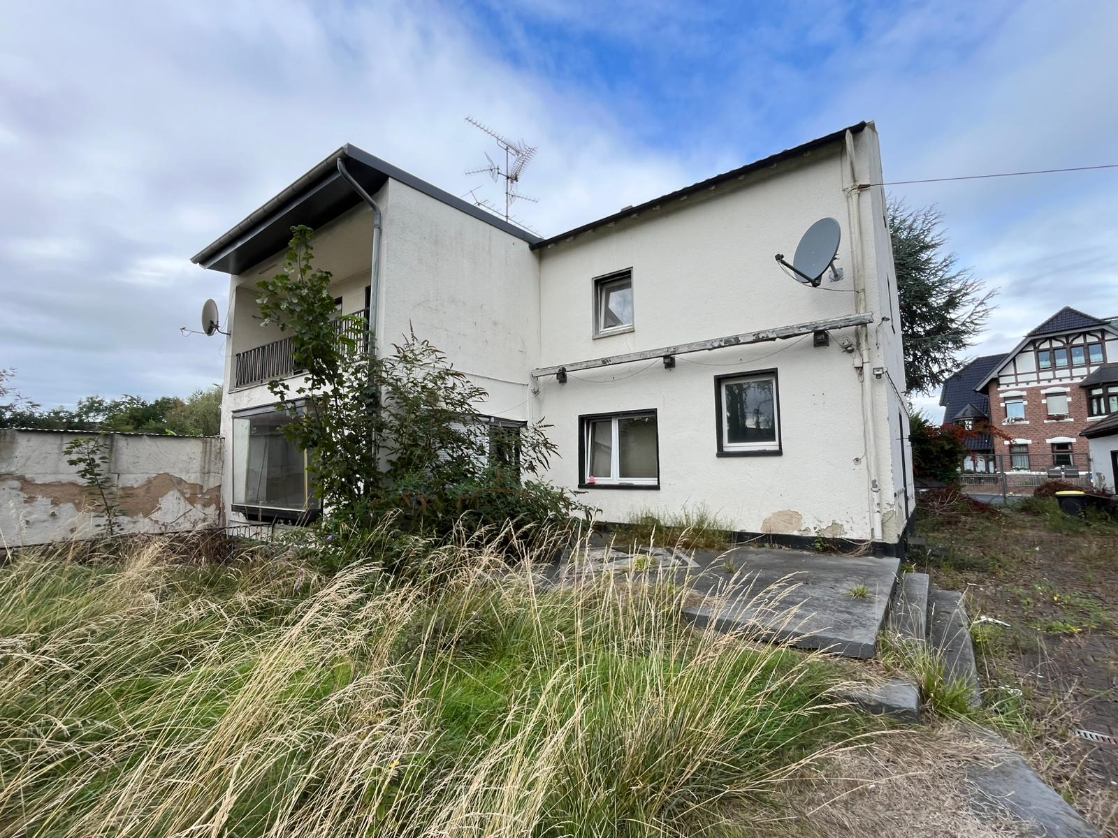 Das Bild zeigt ein zweistöckiges, weiß verputztes Haus. Die Fassade ist teilweise beschädigt. Das Grundstück ist überwuchert mit hohem Gras und Unkraut.