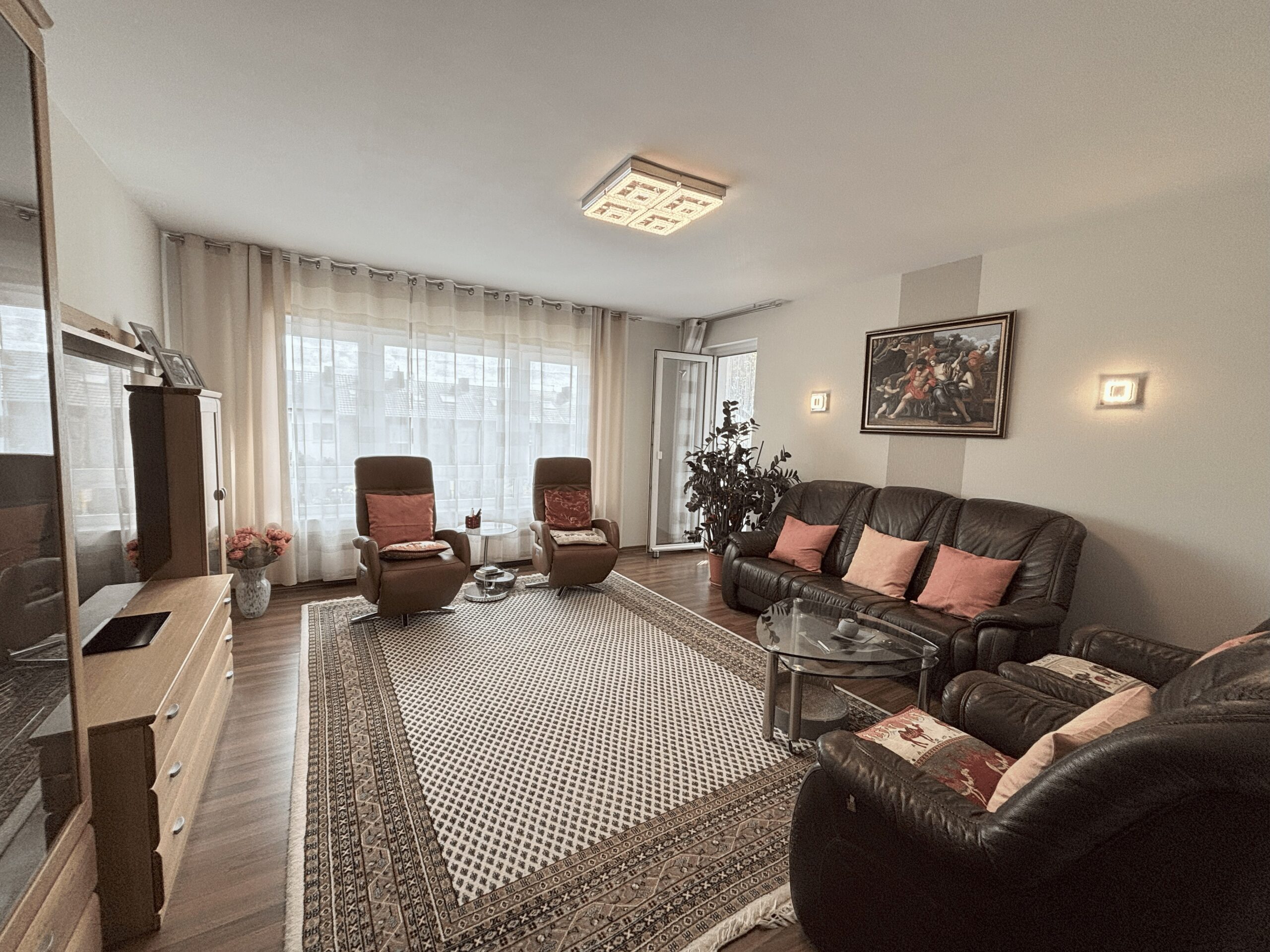 Das Bild zeigt ein Wohnzimmer mit einem großen Fenster, eingerichtet mit Sesseln und einem Sofa, Teppich und einer Schrankwand.